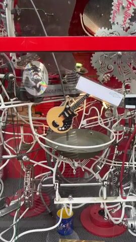 A kinetic sculpture is displayed behind glass. The sculpture is made of various metal objects, including bicycle wheels, gears, a guitar, a CD, and a pot lid. The objects are interconnected by a white wire structure. The background is red. A sign reads "The show must go on". Another sign reads "Touch here to start". A blue and yellow cup is visible at the bottom of the sculpture.