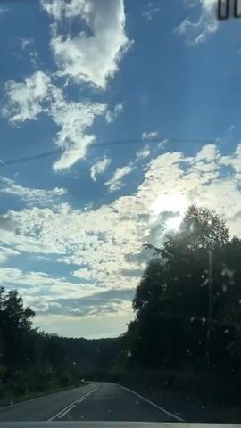 This is a first-person perspective video taken from inside a vehicle, likely a car, traveling on a paved road. The time of day appears to be late afternoon or early evening, as indicated by the sun's position low in the sky, casting bright rays through scattered clouds. The weather is fair with a bright blue sky and fluffy white clouds. The setting is a rural or suburban area with lush green trees lining both sides of the road. The road itself has white lane markings and appears to be a two-lane highway with gentle curves. In the distance, the road continues into a heavily wooded area.

There are no people clearly visible within the frame. The primary "object" is the road and the surrounding natural environment. The overall mood conveyed is one of peaceful travel through a scenic landscape.

Visible text includes a black rectangle with the digits "00" in white, positioned near the top right corner of the frame, likely a timestamp or indicator on the vehicle's dashboard or windshield. Another instance of "00" is visible near the top edge of the frame, potentially part of the same display.