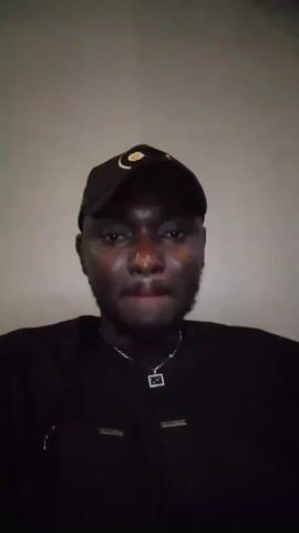 A person with dark skin, wearing a black baseball cap, a black shirt, and a silver necklace with a pendant, is facing the camera. The person looks around and then straightens his head. The background is a light gray wall. The lighting is dim. Sound is audible.