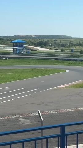 The video shows a go-kart race on an outdoor track on a sunny day. Several go-karts are visible on the asphalt track, which has green grass and trees surrounding it. In the background, there is a blue and white observation tower, and rolling hills with some patchy vegetation. The go-karts are in motion, some in a tight curve, indicating they are actively racing. The perspective is from an elevated position, possibly a spectator area, as evidenced by the railing in the foreground.