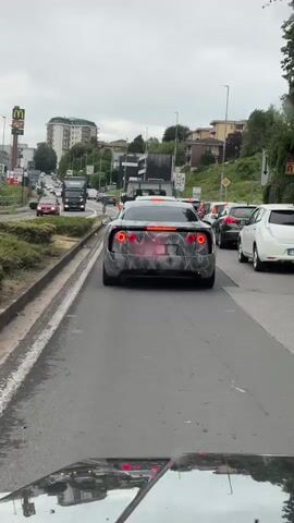 The video shows a camouflaged car driving in traffic. The car is dark with a black and white wavy pattern. The car has red taillights. The license plate is partially visible. There are other cars and trucks on the road. The video appears to be taken from another car.