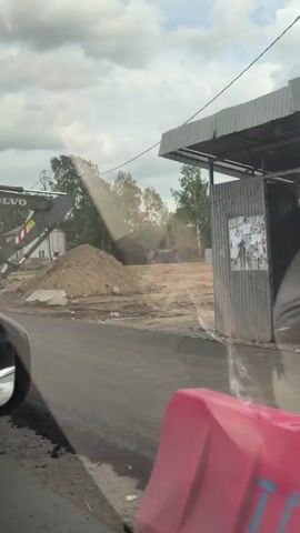 The video shows a Volvo excavator digging into an asphalt road. The excavator is yellow and gray with the words "VOLVO" and "EW205D" visible. The excavator's bucket is digging into the road, leaving a torn-up asphalt surface. There is a pile of dirt in the background. The video is taken from a moving vehicle, and orange and white barriers are visible. The sky is cloudy. Trees and houses are also visible in the background.