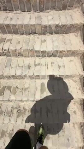 The video shows a first-person perspective of someone walking up a wide stone staircase. The staircase appears to be made of rough, light-colored bricks, worn and somewhat uneven. Sunlight casts distinct shadows on the steps. As the person ascends, the view widens to reveal more of the staircase leading up to a fortified tower. Two people are visible at the top of the stairs, near the tower. The sky above is a bright blue with scattered white clouds. The overall environment suggests an outdoor historical or architectural site.