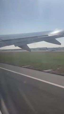 The video captures a view from an airplane window as it takes off or lands on a runway. The airplane wing is prominently visible in the foreground, with its metallic surface reflecting the sunlight. Below the wing, a runway stretches out, with green grass bordering it. In the distance, an airport with terminals and other aircraft can be seen. The sky is clear and blue, with the sun shining brightly. The camera angle suggests a low altitude view, close to the ground, emphasizing the movement and speed associated with air travel. The overall impression is one of transition, either beginning or ending a journey.