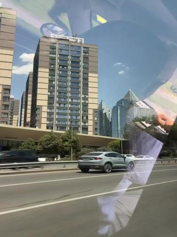 The video captures a view from inside a vehicle, looking out at a cityscape on a sunny day. Multiple cars are visible on a highway, with the closest one being a silver SUV traveling on the right side of the road. Beyond the highway, modern glass skyscrapers with distinctive pointed roofs dominate the background. Trees and greenery are scattered around the buildings. Reflections of the vehicle's interior, including a dashboard and a rearview mirror, are visible on the windshield. The overall environment suggests an urban setting during daylight.