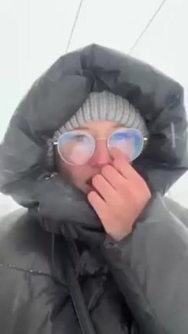 A woman wearing a gray knit hat and round-rimmed glasses is standing in what appears to be a snowy outdoor environment. She is wearing a black puffer jacket with the hood up. The woman is adjusting her glasses with her right hand. The background is blurry, but there appear to be cables overhead. Snow is falling.