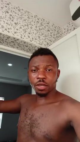 The video shows a shirtless man looking directly at the camera, slightly sweaty. He has short, dark hair with a short, faded haircut. He has a short, groomed beard. Behind him is a doorway leading to another room with grey walls. The ceiling has a white and grey floral pattern. To the right is a white wall with an air conditioner attached near the ceiling. The man speaks directly to the camera, making hand gestures as he talks.