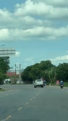 This media file captures a view from a moving vehicle on a paved road in what appears to be a rural or semi-urban area of Lao Bảo Commune, Vietnam. The time of day is daytime, indicated by the bright sunlight and the presence of a blue sky with fluffy white clouds. The weather seems pleasant, with clear skies and a moderate amount of cloud cover.

The primary subjects are two motorcycles and one white pickup truck, all traveling in the same direction on the road. The pickup truck is in the center of the frame, followed by a motorcyclist on the right side of the road and another motorcyclist slightly further ahead and to the right. The road itself is asphalt with yellow dashed lines indicating lanes.

The setting includes lush green trees and vegetation lining the sides of the road. In the background, several tall wind turbines are visible, suggesting the presence of a wind farm or renewable energy infrastructure in the vicinity. Red-tiled roofs of buildings can be seen behind the trees on the left, and utility poles with power lines are also present.

Notable details include traffic signals on the left side of the road and what appear to be signs and trash cans along the right side. The overall atmosphere is one of daily travel or commute, with no strong emotional cues evident. The focus is on the movement of vehicles through the landscape.