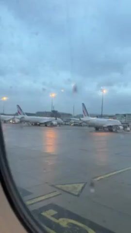 The view from inside an airplane shows a rainy airport scene. Raindrops are visible on the window. Several airplanes, identifiable by the Air France logo on their tails, are parked on the tarmac. Streetlights illuminate the wet ground. The sky is overcast. Yellow ground markings are visible on the tarmac.