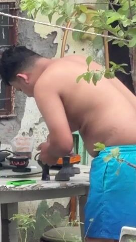 A young man, shirtless and wearing blue swim trunks, stands at a table outdoors, preparing something on a small stove. He is leaning over a clear plastic container filled with food, and he appears to be using a tool to manipulate the contents. A portable stove is visible on the table, as well as other items. The background shows a weathered wall and some greenery. The overall scene suggests a casual, outdoor cooking or preparation activity.