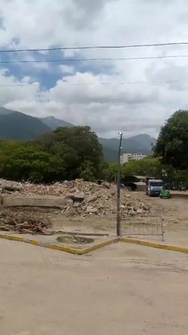 The video shows a scene of destruction with large piles of rubble, likely from collapsed buildings. In the foreground, there is a utility pole with wires and what appears to be a traffic light. In the background, there are mountains and trees, suggesting a natural landscape surrounding the damaged area. A partially collapsed building with a telecommunications tower on its roof is visible to the right. The sky is cloudy. There are no people visible in the video. The overall impression is of a disaster-stricken area.