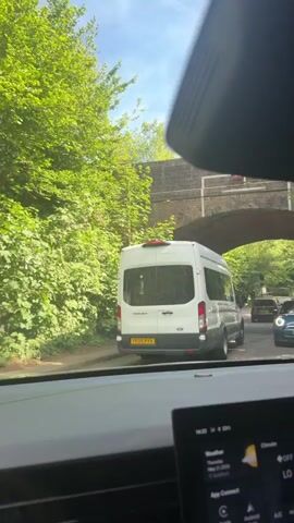 The video shows a car driving under a low-clearance brick bridge. The view is from inside the car, focusing on the road ahead. A white van is driving in front of the car, followed by other vehicles. The road is surrounded by lush green trees. A height restriction sign of "14'0" is visible on the bridge. The sky is blue with some clouds. The car's dashboard is partially visible, showing weather information.