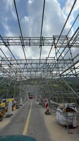 The video shows a vehicle driving on a road under a large scaffolding structure. The scaffolding surrounds the entire road, creating a tunnel-like effect. There are red and white striped traffic cones lining the sides of the road. In the distance, a red car is visible driving in the opposite direction. The sky is partly cloudy, and green hills can be seen in the background.