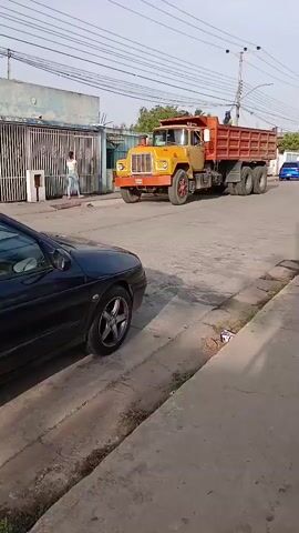 The video shows a yellow and red dump truck moving down a residential street. The truck has two people standing in the back. On the left side of the screen, a black car is parked on the side of the road. A man is walking along the sidewalk on the right side of the street. There are houses and fences on either side of the street, and power lines overhead.  A blue car can be seen further down the street.