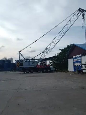 The video shows a landscape scene at what appears to be a port or industrial waterfront area. A large, light-colored crane with a lattice boom dominates the left side of the frame, near a building with signage. Moving further down the line of sight, there are blue cylindrical tanks. In the background, a large ship is docked, painted with white and dark blue stripes. A street light is also visible along the dock. The sky overhead is overcast with patches of sunlight attempting to break through the clouds. The camera appears to be held sideways.