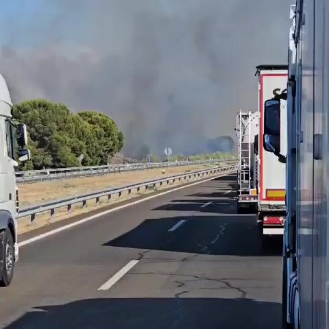 The video shows a highway with several trucks on it. In the distance, there is a large plume of dark smoke rising into the sky, indicating a fire. The trucks appear to be stopped or moving slowly, possibly due to the fire. The landscape around the highway is dry and covered with sparse vegetation. The sky is clear and blue. The video is shot from the perspective of someone in a vehicle, likely one of the trucks.