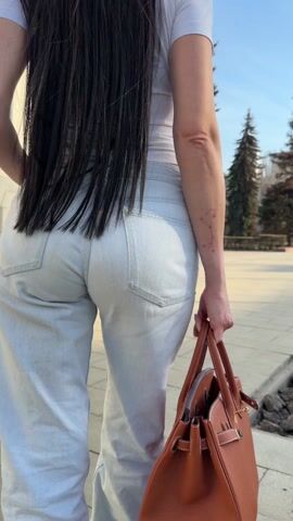 A woman with long, dark hair is walking away from the camera down a sidewalk. She is wearing light-colored jeans, a white t-shirt, and black high-heeled boots. She is carrying a brown leather purse in her right hand. A building with ornate architectural details and several trees are visible in the background. The scene appears to be taking place on a sunny day.
