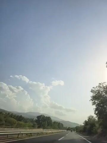 The video captures a first-person perspective of driving on a highway. The scene is dominated by a vast, clear blue sky with a few scattered white clouds on the left. A bright sun is visible on the right side of the frame, casting a soft light. A multi-lane highway stretches ahead, with a metal guardrail on the left side. Trees and green foliage line the sides of the road, with rolling hills and mountains visible in the distance. There are a few cars on the road, all moving in the same direction as the viewer. Speed limit signs are visible on the side of the road. The overall atmosphere is peaceful and scenic, suggesting a journey through a rural or mountainous region.