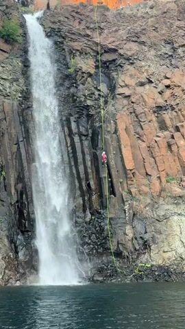 The media file depicts an individual rappelling down a steep, rocky cliff face adjacent to a powerful waterfall. The subject, wearing a red vest with the number "8" visible, is harnessed and secured by a bright yellow rope, indicating they are engaged in a controlled descent. The cliff is characterized by dark, wet rock formations, with streaks of water running down its surface, suggesting recent or ongoing rainfall. Lush green vegetation clings to some parts of the rock, adding a contrast to the rugged terrain. The waterfall itself is a dominant feature, with a torrent of white water crashing down into the dark, rippling water body at the base of the cliff. The overall scene suggests an outdoor adventure activity, possibly caving or waterfall abseiling, taking place in a dramatic natural environment. The lighting appears to be natural daylight, casting some shadows and highlighting the texture of the rocks and the spray from the waterfall. The emotions conveyed are those of focus and determination from the rappeller, set against the raw power and beauty of nature. The location is specified as Talnakh, Russia.
