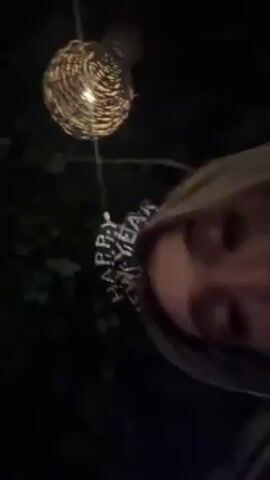 Here's a description of the video:

The video, shot in a low-angle selfie format, appears to be taken at a New Year's Eve celebration at night. The camera focuses on a person wearing a "Happy New Year" headband, glittering with silver letters. Behind the person, the background is dark, with hints of greenery. Above, there's a decorative, round hanging lantern made of woven material, emitting a warm light.  The music playing in the background is upbeat and dance-like. The camera moves around, and at times only the subject's eye is visible in the frame.