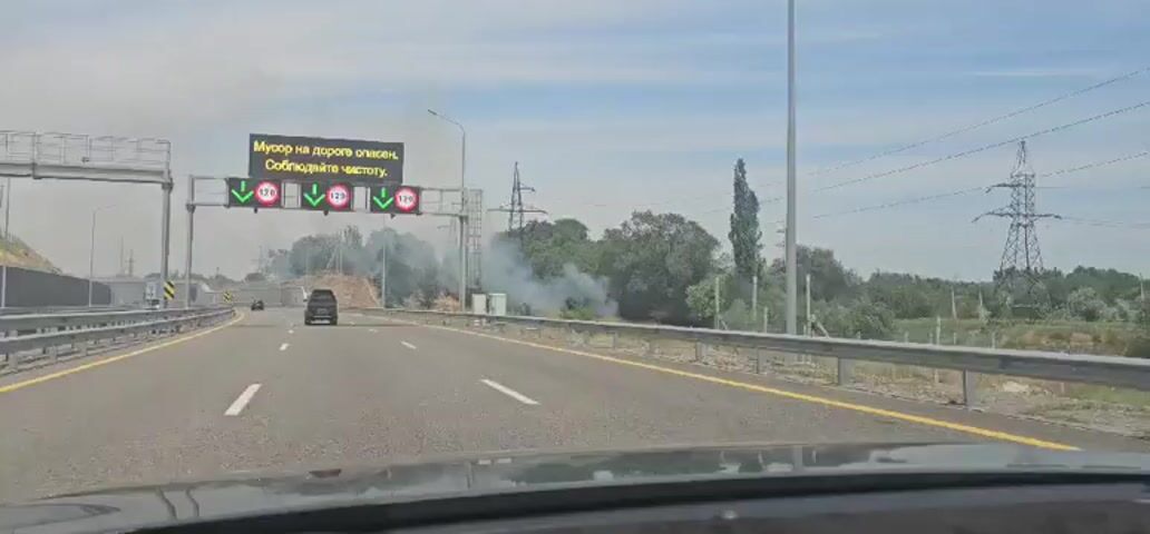 The video shows a view from a moving car on a multi-lane highway. The highway is paved with asphalt and has clear lane markings. In the distance, on the right side of the road, there is a significant amount of smoke rising from vegetation, indicating a fire or controlled burn. Several cars are visible on the highway, including a dark SUV in the foreground.

Above the highway, an electronic billboard displays a message in Russian: "Мусор на дороге опасен. Соблюдайте чистоту." (Trash on the road is dangerous. Keep clean.) Below this text, there are four signs displaying a downward-pointing green arrow and the number "120", likely indicating a speed limit.

The scene appears to be set during the daytime, with a clear blue sky and scattered clouds. The overall mood conveyed is one of caution, given the warning message and the visible smoke, suggesting potential hazards on or near the roadway. The presence of power lines and transmission towers in the background, along with dense green trees and some barren earth, provides context for the roadside environment. The location cues suggest an area that is likely rural or semi-rural, given the open landscape and vegetation alongside the highway.