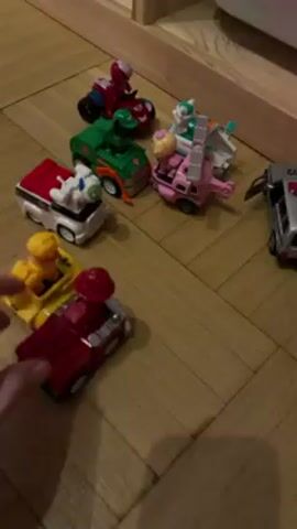 A hand is shown holding a red and grey toy vehicle that has a red figurine on top of it. The hand moves the toy vehicle and puts it back onto the wooden floor. There are several other colorful toy vehicles on the floor as well, including a motorcycle, an SUV, a helicopter, and a construction truck. A metal car is also visible. The audio contains children's voices.