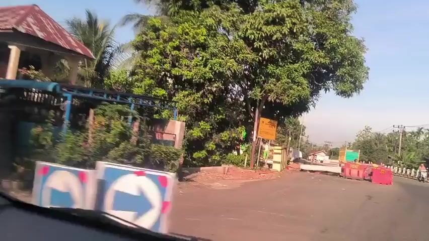From inside a car, the camera captures a view of a road with traffic barriers, a warning sign, and a green-and-white mosque in the background. The mosque has multiple domes. Trees are visible on either side of the road. The sky is clear and blue.