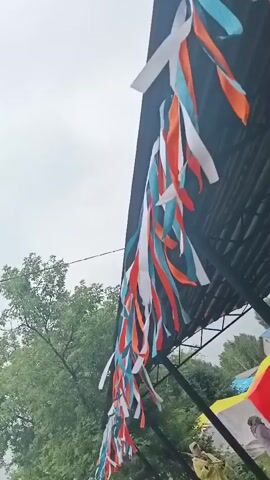 The video shows an outdoor scene, likely a festival or public gathering, in Старые Быги, Russia. The sky is overcast, suggesting a cloudy or possibly rainy day. A black, angular structure, possibly a stage or canopy, dominates the upper portion of the frame. Dangling from its edge is a long string of colorful streamers in white, teal, and orange, creating a festive atmosphere.

In the background, lush green trees are visible, indicating a natural or park-like setting. Below the canopy, there are glimpses of tents or stalls with colorful awnings, including yellow and red. A few individuals can be seen, though they are small in the frame and their activities are not clearly discernible. One person in the lower right appears to be wearing a light-colored outfit. The overall impression is one of an outdoor event underway, with decorations indicating a celebration. The weather seems to be damp or cool, given the overcast sky.