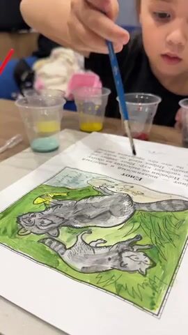 A young girl is painting a picture of two raccoons on a piece of paper. She is holding a paintbrush in her right hand and is focused on her work. There are several plastic cups filled with different colors of paint in the background, suggesting she is using watercolors. The drawing depicts the raccoons in a grassy area. The girl is wearing a black shirt. The scene appears to be indoors, possibly at a table, with other painting supplies visible. The lighting suggests it could be daytime. The girl's expression is one of concentration. There is Cyrillic text visible on the page, above the drawing.