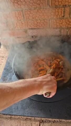 A person in a kitchen stirs a large pot of food on a stovetop. The food is dark brown with orange strips of carrot and steams in the cast iron pot. The person's right arm and hand hold a metal tool to stir the food in the pot. The person is wearing a short-sleeved brown shirt. The stovetop is black, and the wall behind it is made of textured, light orange bricks. The lighting is bright and even.
