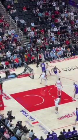 The video shows a basketball game in progress. Players in white jerseys with red trim are on offense, while players in purple and gold jerseys are on defense. The court has a large red section in the center. The camera angle is high, providing an overview of the court and a section of the audience. Many spectators are in the stands, mostly dressed in casual attire. A referee is on the court, monitoring the play. The player in purple drives to the basket and falls to the ground as he tries to score.
