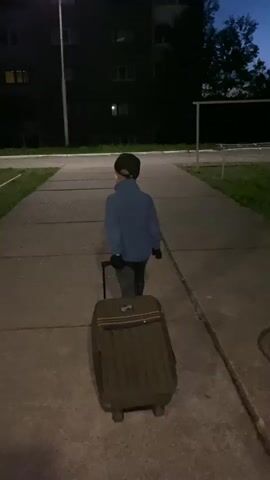 A boy is walking away from the camera, pulling a suitcase along a paved walkway at night. He is wearing a blue jacket, dark pants, and a baseball cap. The walkway is bordered by grass and a building with illuminated windows is visible in the background. A streetlight illuminates the scene, casting a shadow of the boy and his suitcase on the pavement. A soccer goal is also visible in the background.