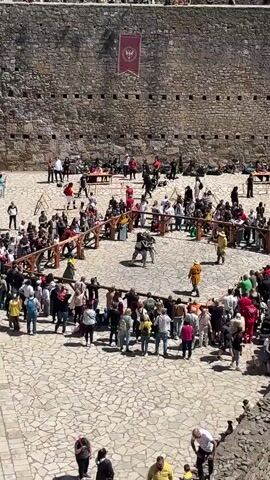 The video shows a jousting event taking place in a stone courtyard. A crowd surrounds a wooden barrier forming a ring where two participants in full armor engage in combat using swords. The participants are facing each other and striking their swords together. The crowd consists of people of various ages and clothing styles, many of whom are holding up their phones to record the event. In the background is a high stone wall with small windows and a banner hanging near the top. Additional people are scattered in the background near tables and other areas of the courtyard. The weather appears to be sunny and clear.