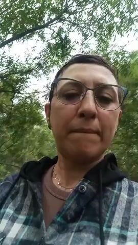 A medium shot shows a person wearing glasses and a plaid jacket in a forest. The person has short, dark hair. The background consists of trees, including birch trees, and green foliage. The lighting is natural, suggesting an outdoor setting during the day. The camera appears to be held by the person, indicating a selfie-style recording. The person is looking directly at the camera.
