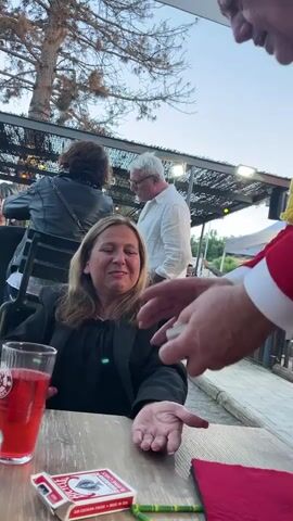 The video shows a woman sitting at a wooden table with a deck of cards and a glass of red liquid in front of her. She is looking up and smiling. A man in a red jacket with yellow trim is standing over her, holding cards. He appears to be performing a magic trick, showing her two cards. In the background, other people are visible, and there is a covered outdoor seating area with lights.