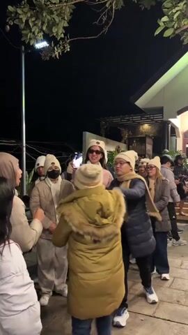 A group of people, most wearing white beanies and puffer jackets, are gathered outdoors at night. One person in the center is holding a smartphone in landscape orientation, appearing to take a photo or video of the group. The person holding the phone is wearing a pink puffer jacket and light grey pants. To their right, a person wearing a yellow puffer jacket and a tan scarf is looking at the phone and making a peace sign with their fingers. Other individuals are visible behind them, some also looking towards the phone. The background includes a building with lights and some trees. The ground appears to be paved with stone.