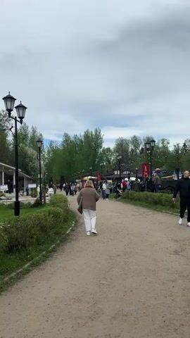 The video shows a dirt path in a park on an overcast day. Along the path are several people walking in both directions. There are lampposts lining the path, and green bushes are growing on either side. In the distance, there appears to be a gathering of people with decorations and possibly some vehicles. There are trees visible in the background. The overall scene suggests an outdoor event or fair in a park setting.