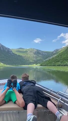 Two young boys are lying face down on the deck of a boat, looking out at a picturesque natural landscape. The boat appears to be moving on a calm body of water, likely a lake or river, bordered by lush green, tree-covered mountains under a clear blue sky with scattered white clouds. The scene suggests a peaceful and scenic outing. The boy on the left is wearing a blue t-shirt and green shorts, with his feet up and visible. The boy on the right is wearing a dark hoodie and black shorts, with his legs stretched out. The overall mood is one of relaxation and enjoyment of the natural beauty. The time of day appears to be daytime, likely late morning or early afternoon, given the bright sunlight and the shadows. There is no visible text in the media file.