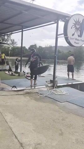 A wake park is shown with a body of water, a dock, and a covered area. There are several people present. One person in a wetsuit walks with a wakeboard towards the water. Another person stands on the dock, shirtless. A person in the water is holding onto a flotation device. Inside the covered area, people are sitting and watching. Wakeboards are visible on the dock. Trees and hills are in the background.