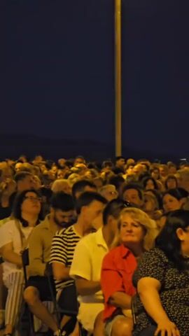 A crowd of people are sitting and standing outdoors at night. The sky is dark blue. In the background, there is a large industrial facility with many lights and tall structures, including a prominent smokestack. A woman and a young boy are walking towards the camera in the foreground. The woman is wearing a black tank top and a dark skirt, and the boy is wearing a black t-shirt with a yellow logo. They appear to be interacting. Other people in the crowd are looking in various directions, some towards the industrial facility and others elsewhere. The overall atmosphere seems to be one of observation or attendance at an event.