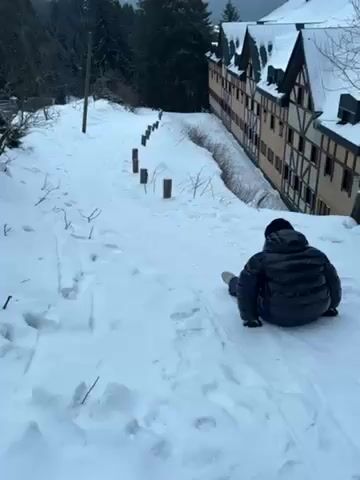 A person wearing a black puffer jacket, black gloves, and a black hat is sitting in the snow on a snowy hillside. They appear to be attempting to slide down the hill on their bottom. The hill is covered in snow and has some tracks already in it. There are wooden posts lining the side of the hill. To the right of the hill is a large building with a snow-covered roof. Behind the hill are trees and a cloudy sky. The person is moving slowly down the hill.