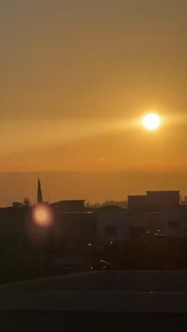 This is a vertical, handheld video clip capturing a sunset over a town in Katzrin, Israel. The sun is a bright, luminous orb positioned in the upper right quadrant of the frame, casting a warm, golden glow across the sky. A soft haze or thin clouds streak horizontally across the sky, softening the sun's intense light.

In the mid-ground, a silhouetted cityscape is visible. Buildings with flat roofs and rectangular structures dominate the scene, with some appearing to be multi-story. A tall, slender minaret-like structure rises above the rooftops on the left side of the frame, suggesting a religious building. Trees are sparsely scattered amongst the buildings. A prominent lens flare, appearing as a reddish-pink circular glow, is situated in the lower left quadrant, partially obscuring some of the buildings. In the far distance, faint, layered mountain ranges are visible against the horizon.

The foreground shows a dark, horizontal surface, possibly a rooftop or balcony, from which the video is being filmed. The overall mood is serene and tranquil, typical of a sunset. The time of day is clearly dusk, as indicated by the setting sun and the warm, orange hues of the sky. The weather appears clear, with only a light haze present. No people are visible in the frame. There is no visible text in the media.