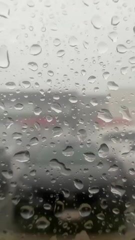 This is a close-up, vertical shot of water droplets on a glass surface, likely a window. The background is out of focus, showing a blurred scene with indistinct shapes of buildings and vehicles. The lighting suggests an overcast or dim day. The water droplets vary in size and shape, some appearing round and clear, others stretched and elongated, indicating movement or condensation. The overall impression is one of a rainy or misty environment.