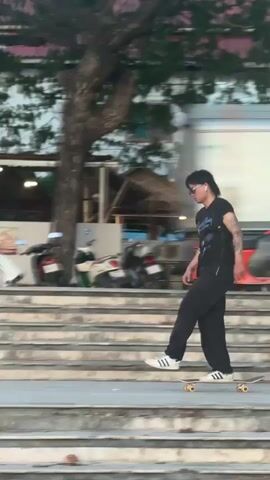 A young person with dark hair and sunglasses is skateboarding down a set of stairs. They are wearing a black t-shirt, black pants, and white sneakers. The skateboard is white with black wheels and has three stripes on the side. In the background, there are buildings, trees, and a black car. Another person wearing an orange t-shirt is walking up the stairs. The scene appears to be outdoors, possibly on a street or promenade.