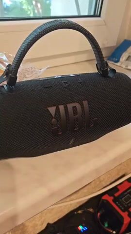 A black JBL Xtreme 3 portable speaker with a dark gray fabric handle is placed on a windowsill. The speaker is oriented horizontally, with the JBL logo prominently displayed in black with a subtle red outline. The speaker's fabric mesh is visible, as are the '+' and '-' volume buttons on top. The handle is attached to the speaker with black clips. In the background, a window shows a blurred view of greenery and what appears to be a building, suggesting an indoor setting. Below the speaker, a small, red electronic device with colorful LEDs is visible, though its function is unclear. The overall lighting suggests it is daytime, possibly late morning or afternoon. The scene is static, focusing on the object. No people are visible. There is no immediate indication of weather conditions, but the greenery suggests it is not winter. The provided location context is Odesa, Ukraine.
