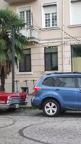 The video shows a red convertible classic car parked on a cobblestone street next to a blue SUV. Behind the cars, a palm tree stands tall, its fronds fanning out against a building. The building has several windows with decorative grills and a balcony. Two electric scooters are parked near the building. The video begins by showing the rear of the red car and the side of the blue car, then pans to the left, revealing more of the red car's exterior and the building. The camera continues to pan, showcasing the full side profile of the red convertible and the surrounding environment.