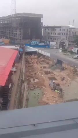 The video shows an aerial view of a construction site with several buildings in the background. The foreground features a large excavation area filled with mud and water, where several workers are present, engaged in what appears to be construction work. Some workers are seen in the excavation pit, while others are on the perimeter. There are piles of dirt and debris scattered around the site. A partially constructed building with scaffolding is visible to the left, and another building, seemingly completed, is to the right. The sky is overcast, suggesting it might be a cloudy day.
