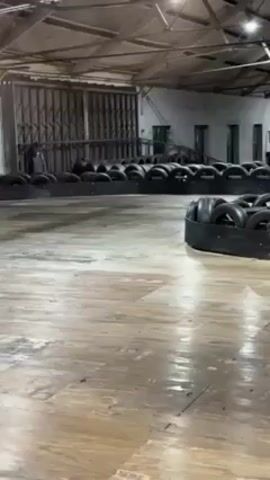 The video shows three go-karts racing on an indoor track. Each driver wears a helmet and dark clothing, and each kart is predominantly blue and black. The track is defined by black tires that form the boundary. The flooring appears to be a wooden surface with visible wear and tear. The indoor space is large and warehouse-like, with a high ceiling, exposed beams, and large metal doors on one side. The lighting appears to be a mix of natural light coming through the doors and artificial lighting fixtures in the ceiling.