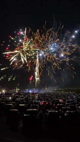 The video shows a nighttime fireworks display. Numerous cars are parked in rows, facing away from the camera, creating a vast parking lot filled with spectators. Above the cars, fireworks illuminate the dark sky with bursts of light in various colors including red, white, gold, and blue. Some fireworks are large, explosive bursts, while others are streaks of light shooting upwards. In the background, faint lights suggest a distant urban area or event venue. The scene is one of communal viewing of a pyrotechnic event.