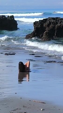 A woman with dark hair lies prone in shallow water on a beach. The wet sand reflects her body. Waves lap at the shore, and large, dark rocks jut out from the sea on either side of her. Further out, the blue ocean stretches towards the horizon, where a single surfer can be seen riding a wave. A stick lies on the sand in the foreground.